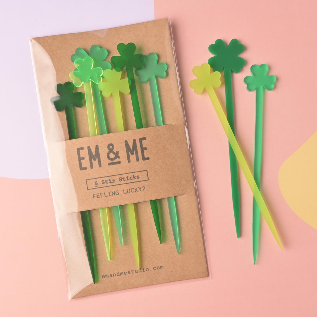 A packaged set of EM & ME 'Feeling Lucky?' clover-tipped acrylic stir sticks (6 pieces in multiple shades of green) in a clear sleeve with a kraft paper backing. Three loose clover stir sticks (dark, medium, and lime green) are displayed beside the package on a pink and yellow background.