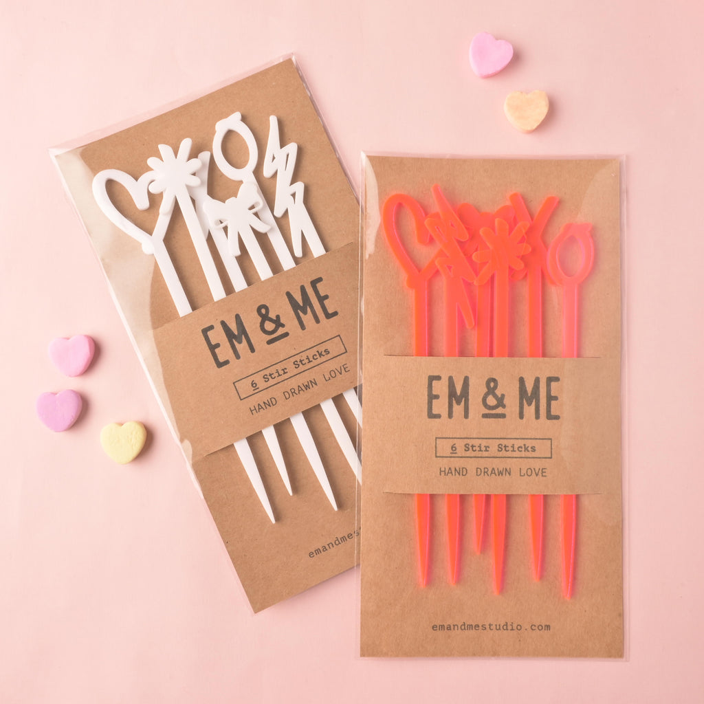 Two packaged sets of EM & ME '6 Stir Sticks Hand Drawn Love' acrylic drink stirrers. The left set is white and the right set is fluorescent pink, featuring various novelty shapes (hearts, lightning bolts, bows, etc.). Conversation heart candies are scattered nearby on the pink background.