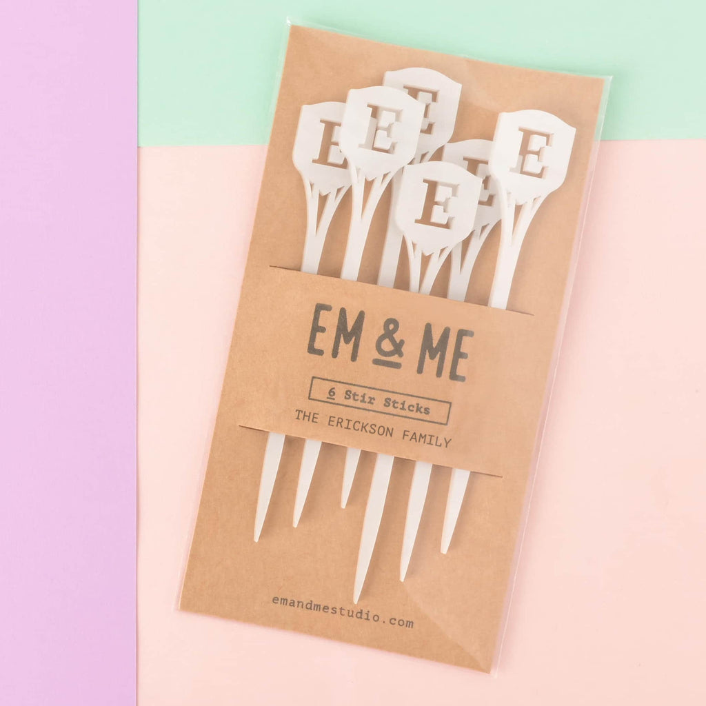 A packaged set of six white acrylic stir sticks from EM & ME, featuring a shield or plaque design topped with the initial 'E' (The Erickson Family). The sticks are packaged in a clear sleeve with a kraft paper backing card. Personalized Family Shield Stir Sticks, Family Letter Initial Swizzle Sticks, Thanksgiving Bar cart, Stocking Stuffer