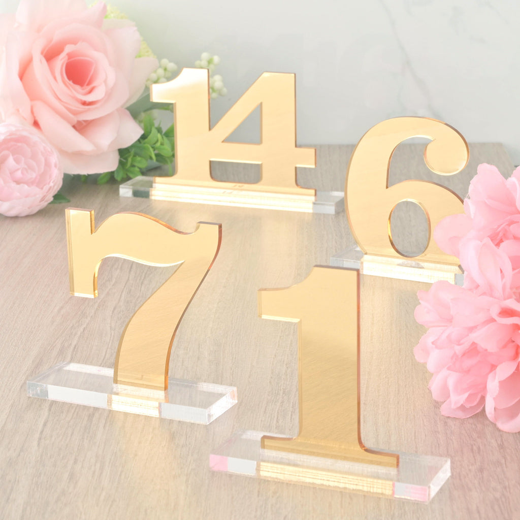 A collection of four gold mirrored acrylic table numbers (1, 6, 7, and 14) with clear acrylic bases, used as elegant event décor and set against soft pink flowers and a wooden surface.