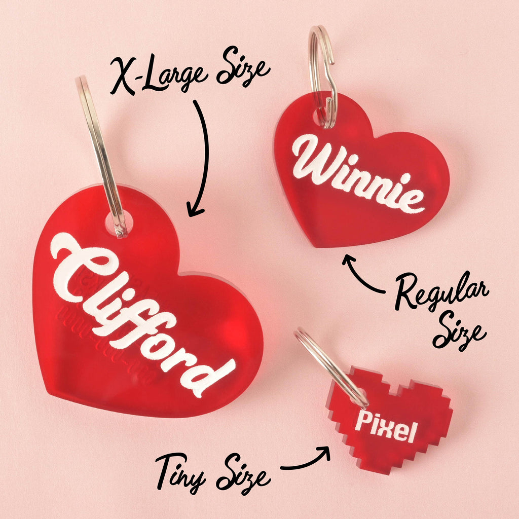 A product photo showing three red, heart-shaped pet ID tags. The largest, labeled "X-Large Size," has "Clifford" engraved on it. The middle tag, labeled "Regular Size," has "Winnie" engraved on it. The smallest tag, labeled "Tiny Size," is a pixelated heart shape with "Pixel" engraved on it. All tags are shown with a silver split key ring attachment.