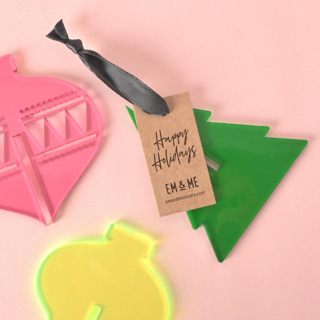 Flat lay of colorful acrylic Christmas ornaments—a pink bauble, neon yellow bauble, and green tree—with black ribbon and a kraft “Happy Holidays” brand tag on a light pink background.
