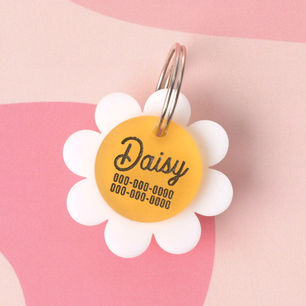 Custom daisy-shaped pet ID tag made of white and yellow acrylic, featuring the name Daisy and two contact phone numbers in black lettering, shown on a pink abstract background