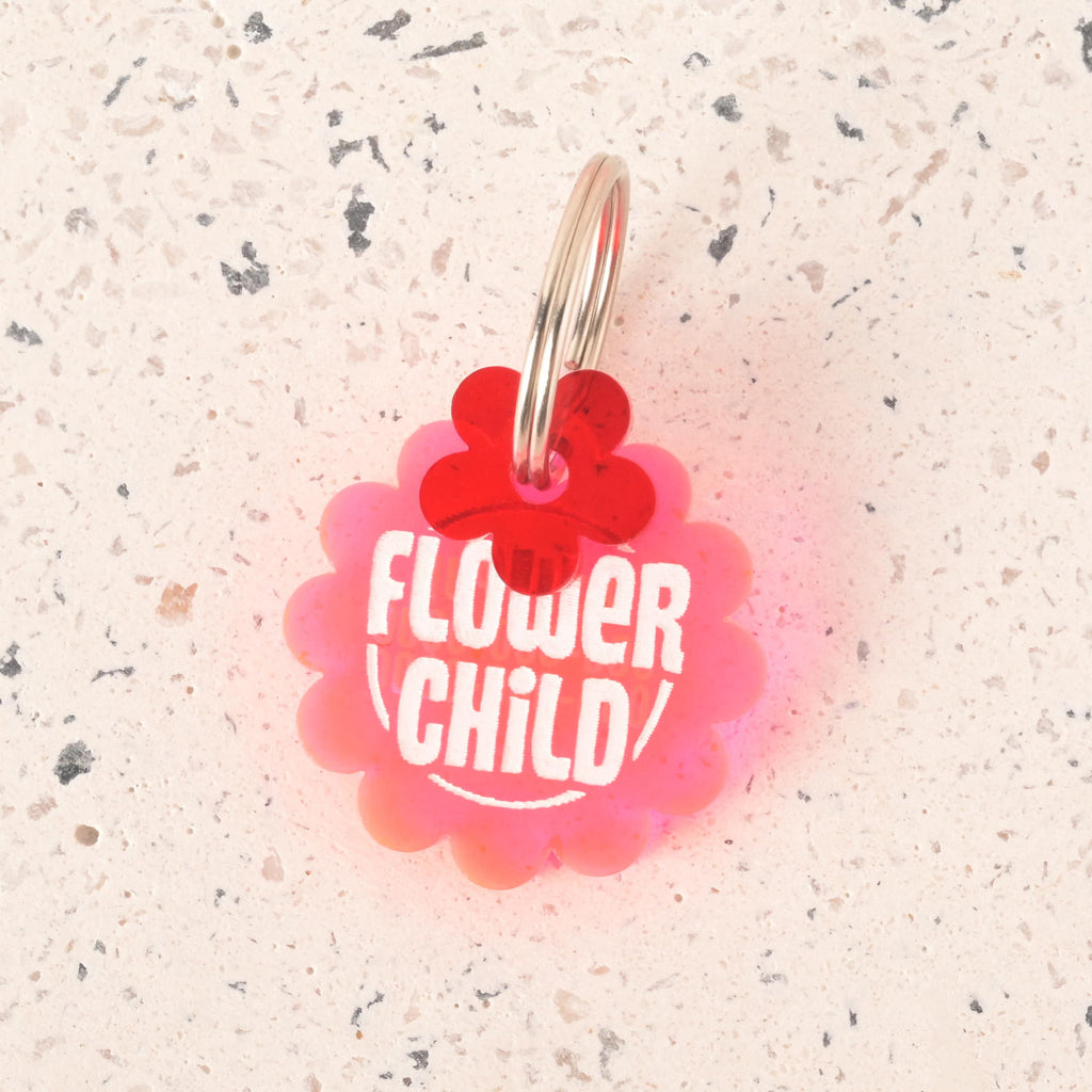 A pink, flower-shaped pet tag that says "FLOWER CHILD" in white letters. A small red flower charm is attached to the tag. Both are connected to a silver ring, perfect for a collar. The items are shown on a light-colored, speckled surface.