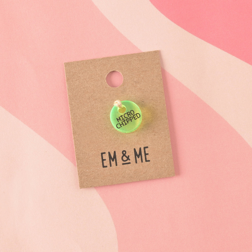 A small, circular, fluorescent neon green acrylic charm engraved with "MICROCHIPPED" in black text. The charm is secured with twine to a branded 'EM & ME' kraft card.