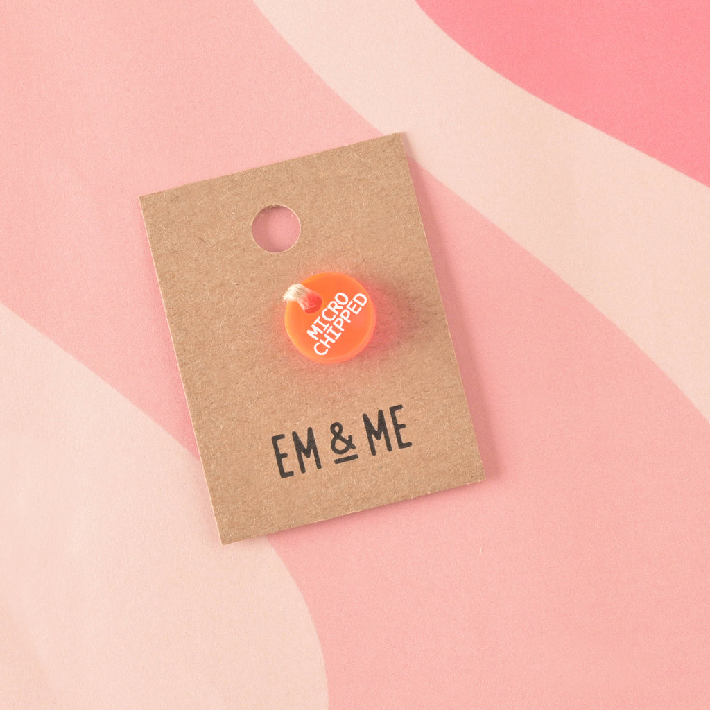 A small, circular, fluorescent orange acrylic charm engraved with "MICROCHIPPED" in white text. The charm is secured with twine to a branded 'EM & ME' kraft card.