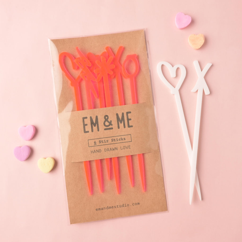A packaged set of EM & ME translucent orange acrylic 'Hand Drawn Love' stir sticks (6 pieces) in a clear sleeve with a kraft paper backing. Two loose white acrylic stir sticks (a heart and an XO shape) and scattered pastel conversation heart candies are displayed nearby on a pink background.