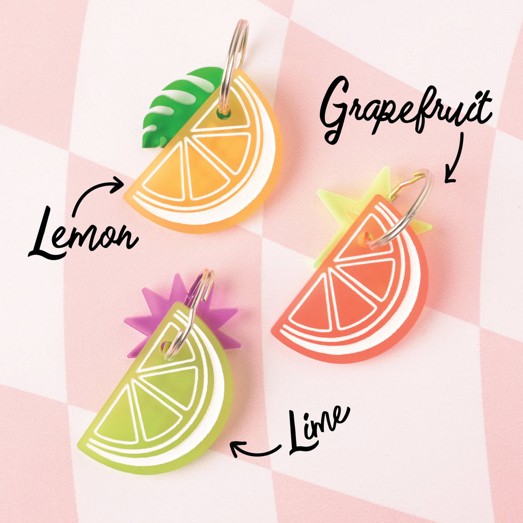 A set of three acrylic citrus slice charms: a yellow lemon with a green leaf charm, a green lime with a pink sunburst charm, and a red-orange grapefruit with a yellow star charm. Each is a layered accessory shown on a checkered background.