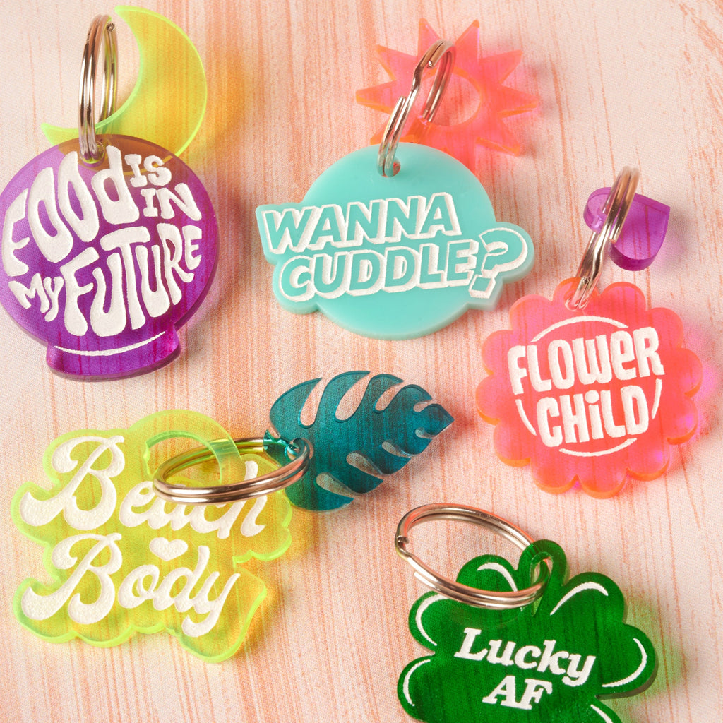 A collection of six colorful acrylic novelty tags/keychains with bold text slogans. Tags include: a purple crystal ball shape ('Food Is In My Future'), a blue bubble shape ('Wanna Cuddle?'), a pink flower shape ('Flower Child'), a lime green wavy shape ('Beach Body'), a green clover shape ('Lucky AF'), and a blue Monstera leaf charm. Lucky AF, 4 leaf Clover Personalized Pet Tag, St Patrick Cat and Dog ID Tag