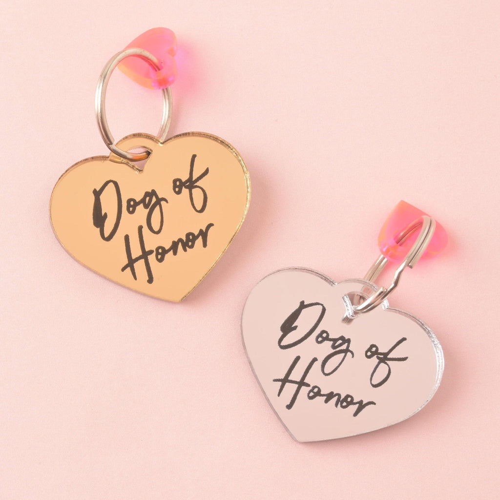 Two personalized heart-shaped pet ID tags, both engraved with the script text "Dog of Honor." One is mirrored gold and the other is mirrored silver acrylic. Both tags are accented with a small iridescent pink heart charm and displayed on a pink background.