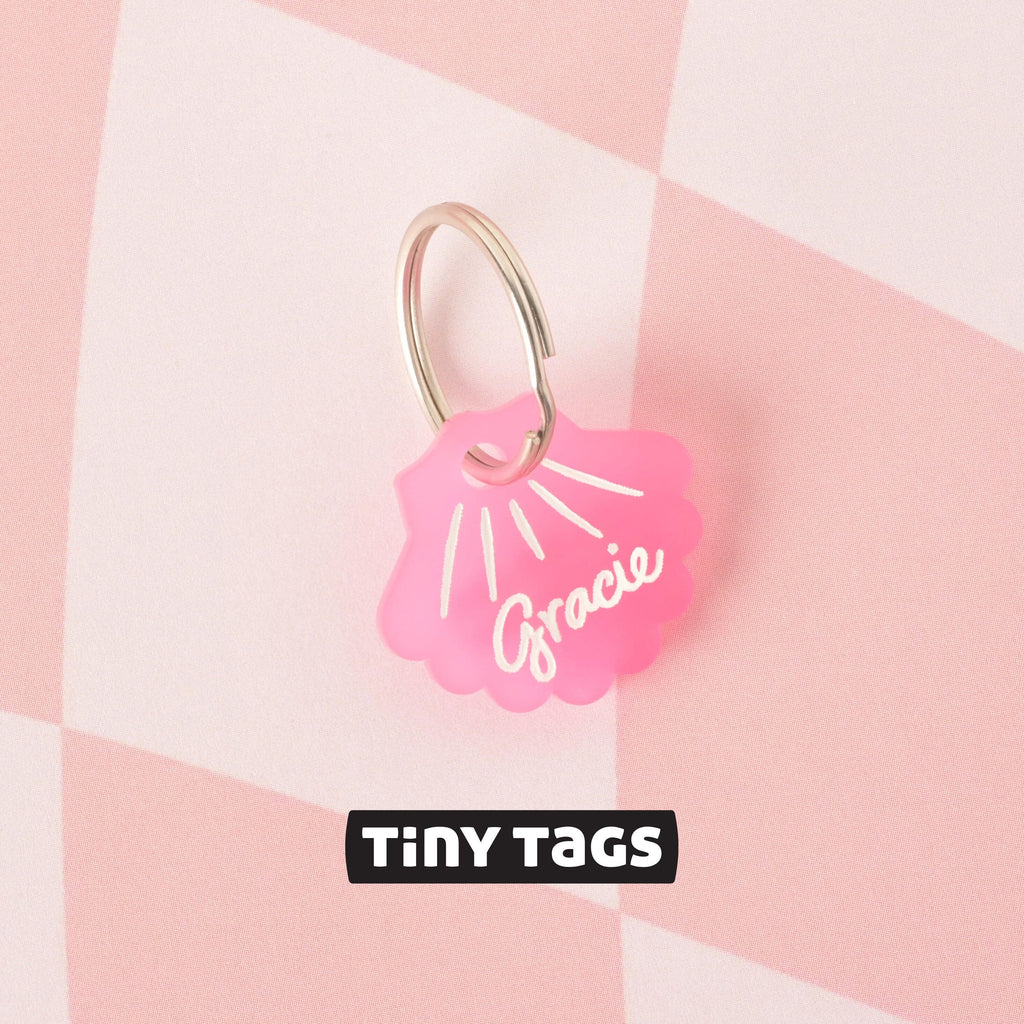 A close-up shot of a small, pink seashell-shaped pet tag that says "Gracie," attached to a silver ring. The tag is resting on a pink and white checkered surface. The image also contains the text "TINY TAGS" at the bottom.