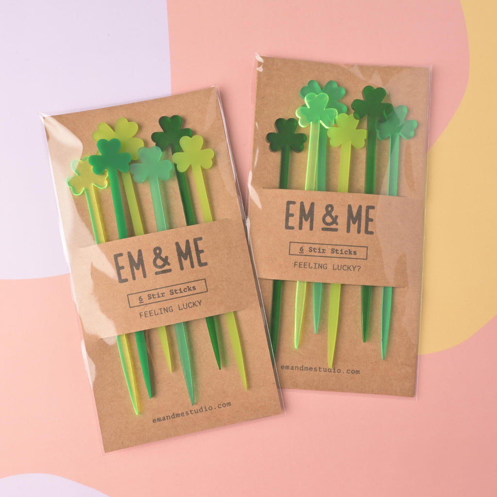 Two packaged sets of EM & ME 'Feeling Lucky?' clover-tipped acrylic stir sticks. Each package contains 6 sticks in various shades of green (lime, medium, and dark), displayed in clear sleeves with a kraft paper backing card.
