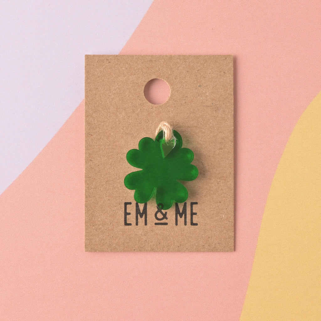 A single translucent dark green acrylic charm shaped like a four-leaf clover, displayed on a small square kraft paper backing card with the EM & ME logo printed below it. The card has a circular hole at the top and is set against a color-blocked background.