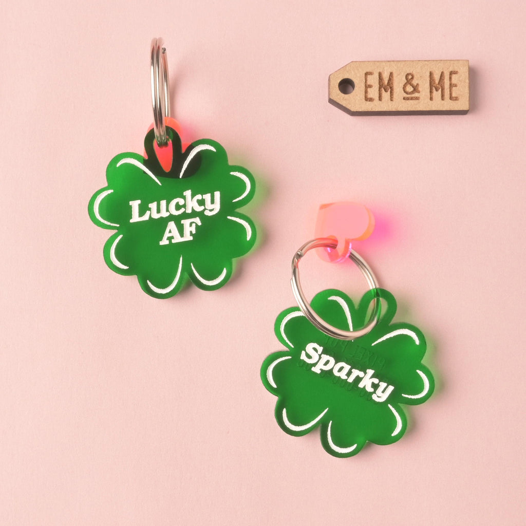 Two dark green, clover-shaped acrylic pet ID tags on a pink background. The top tag features the slogan 'Lucky AF,' and the bottom tag is personalized with the name 'Sparky.' Both have small pink charms attached. A small wooden EM & ME brand tag is visible in the top right corner. Lucky AF, 4 leaf Clover Personalized Pet Tag, St Patrick Cat and Dog ID Tag