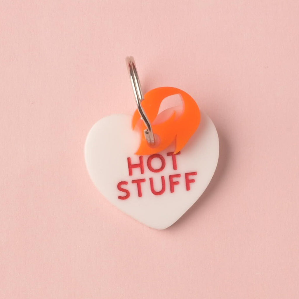 Close-up of a white heart-shaped acrylic pet ID tag, engraved with the red slogan 'HOT STUFF.' An orange flame charm is layered and attached to the silver split ring, displayed on a pink background. Conversation Hearts, Valentines Heart Dog tag, Customize front and back, ID tag for cats and dogs, Personalized Pet Tag