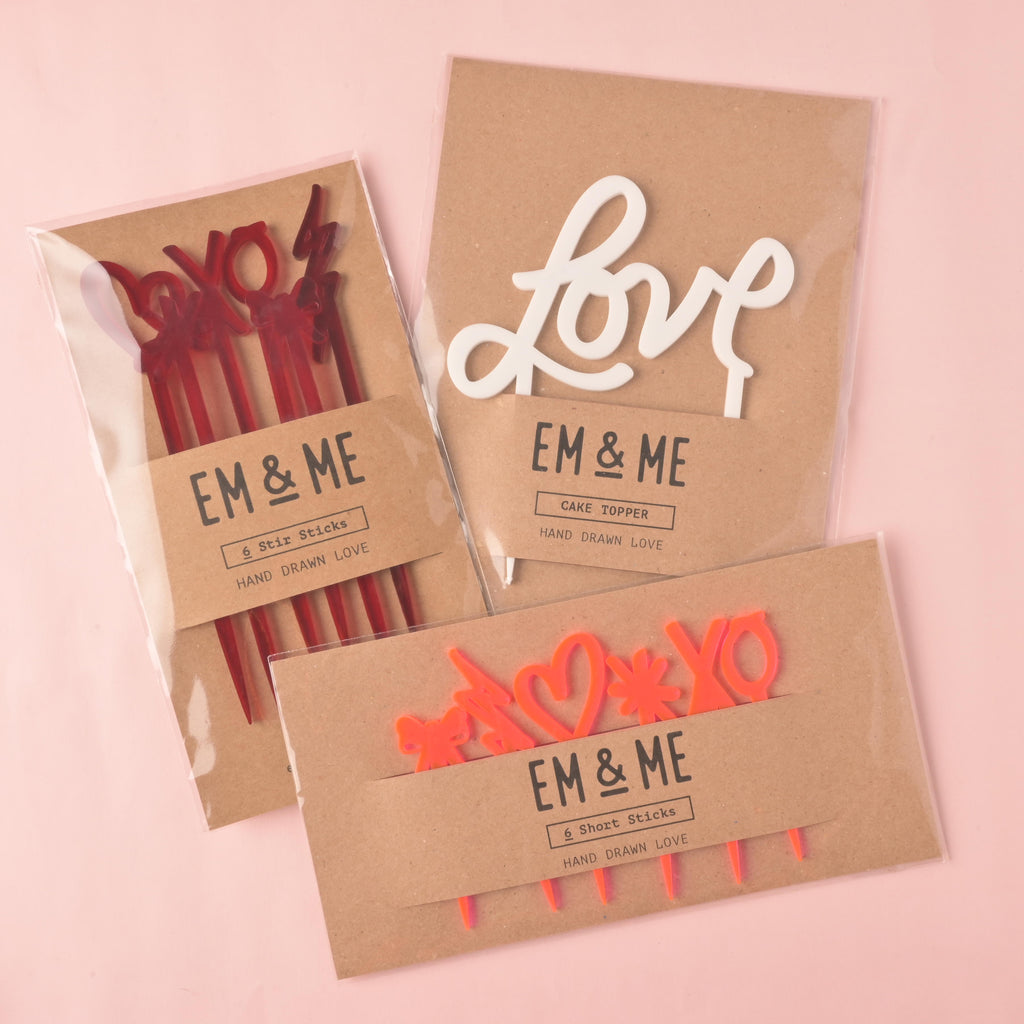 Three EM & ME products packaged in clear sleeves with kraft paper backing, displayed on a pink background. Products include a white 'love' script cake topper, a set of red 'XO' and lightning bolt stir sticks, and a set of orange 'Shoe' stir sticks featuring hearts and other shapes.
