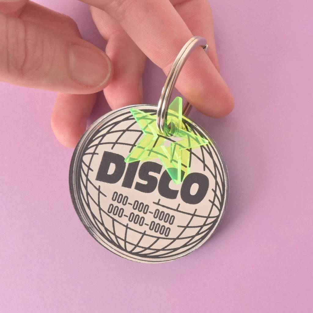 A person holds a large, personalized circular disco ball-shaped pet tag. The tag has the word "DISCO" and a placeholder phone number on it. A small, translucent green star charm is attached. The items are shown against a solid purple background.