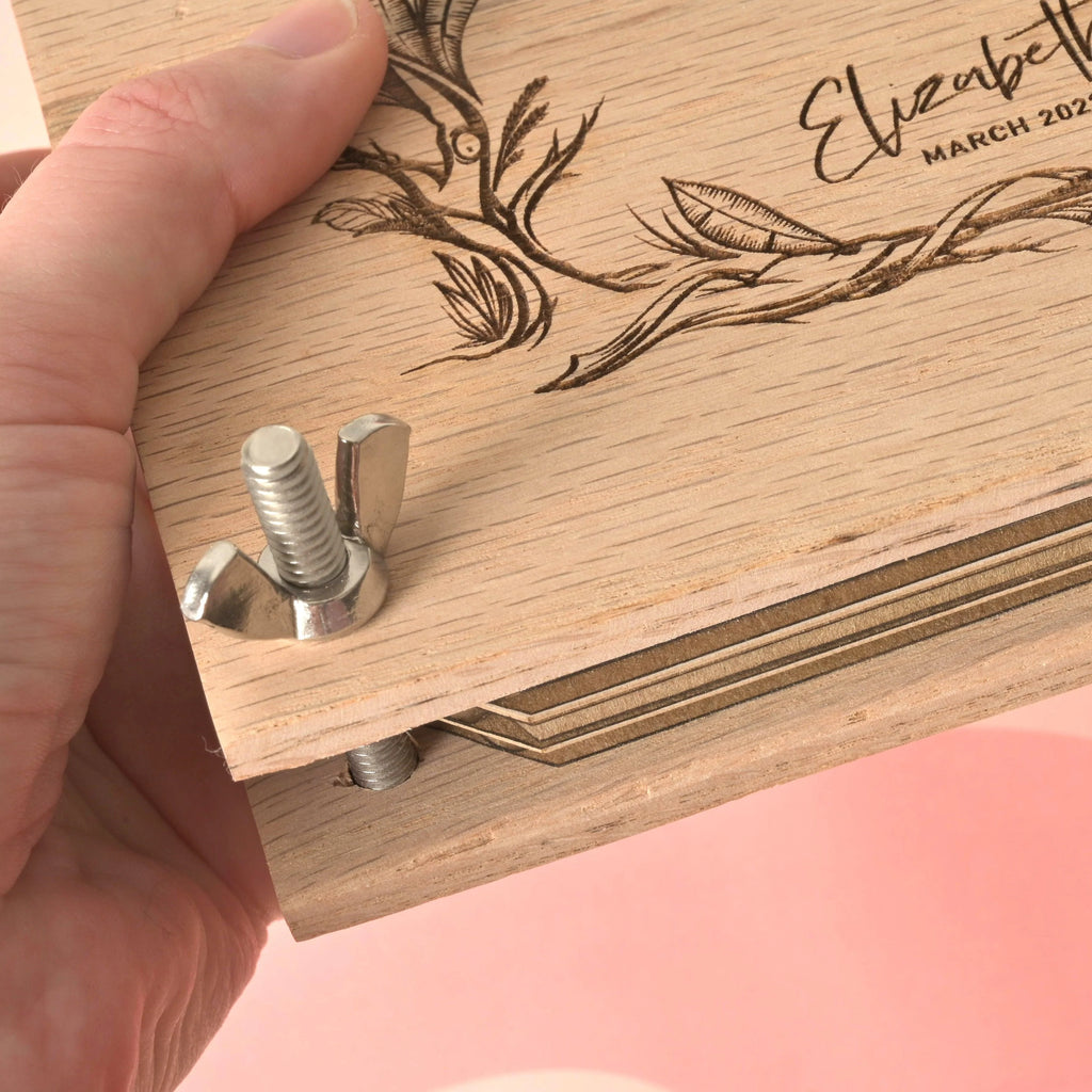 Close-up of a hand tightening screw on oak flower press kit with engraved floral design and cardboard layers inside