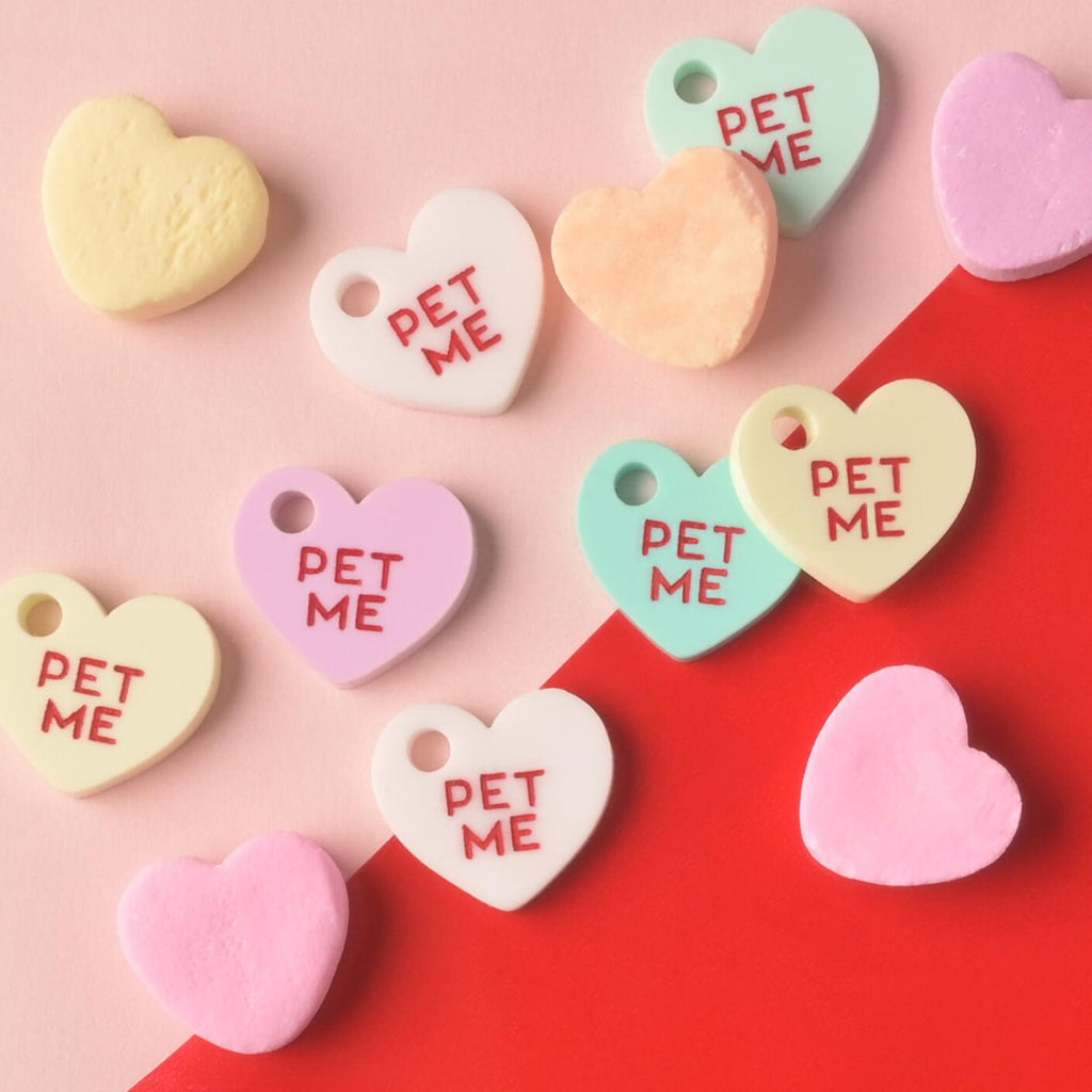 An arrangement of several heart-shaped acrylic pet charms, each engraved with the red slogan 'PET ME,' mixed among actual pastel conversation heart candies. The colors include white, pink, mint green, and yellow, displayed on a color-blocked pink and red background. Small Pet Me Heart, Candy Inspired, Valentine's Day, Seasonal Mini, Tiny Accessory for Pet ID Tag, Cats and Dogs Add-on Charm