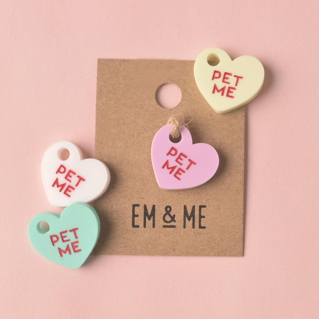 Four small, heart-shaped acrylic mini-charms, each engraved with the red slogan 'PET ME.' The charms are in pastel colors (pink, white, mint green, and yellow). One pink charm is attached to an EM & ME branded kraft paper backing card. Small Pet Me Heart, Candy Inspired, Valentine's Day, Seasonal Mini, Tiny Accessory for Pet ID Tag, Cats and Dogs Add-on Charm