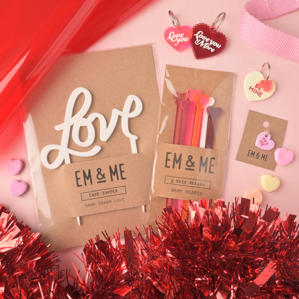 A flat lay of EM & ME Valentine's Day products on a pink background, accented with red tinsel and conversation heart candies. Products shown include a white 'love' script cake topper, packaged heart stir sticks, and several heart-shaped pet tags with phrases like "Love You" and "Be Mine."