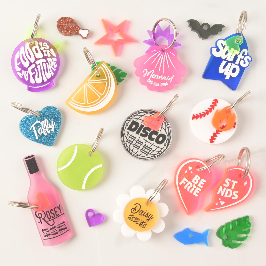 A large collection of colorful, personalized pet tags and charms scattered on a white surface. The tags come in various shapes and phrases, including "FOOD IS IN MY FUTURE," "Surf's Up," "Taffy," "Mermaid," "Disco," "Rosey," "Daisy," and "Best Friends" heart halves. Charms include an orange slice, a tennis ball, a baseball, a wine bottle, and small accents like a glittery drumstick, bat, and heart. Each tag is attached to a silver ring for a collar.