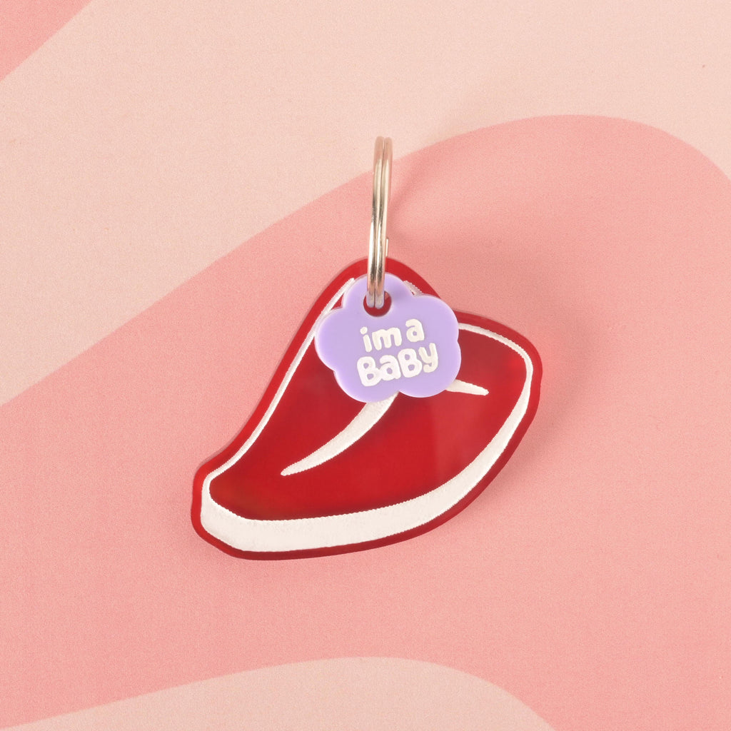 A red steak-shaped pet ID tag with a small purple cloud charm that says "i'm a Baby" in white letters. The tag is attached to a silver ring, perfect for a collar, and is displayed on a pink background with a wavy design.