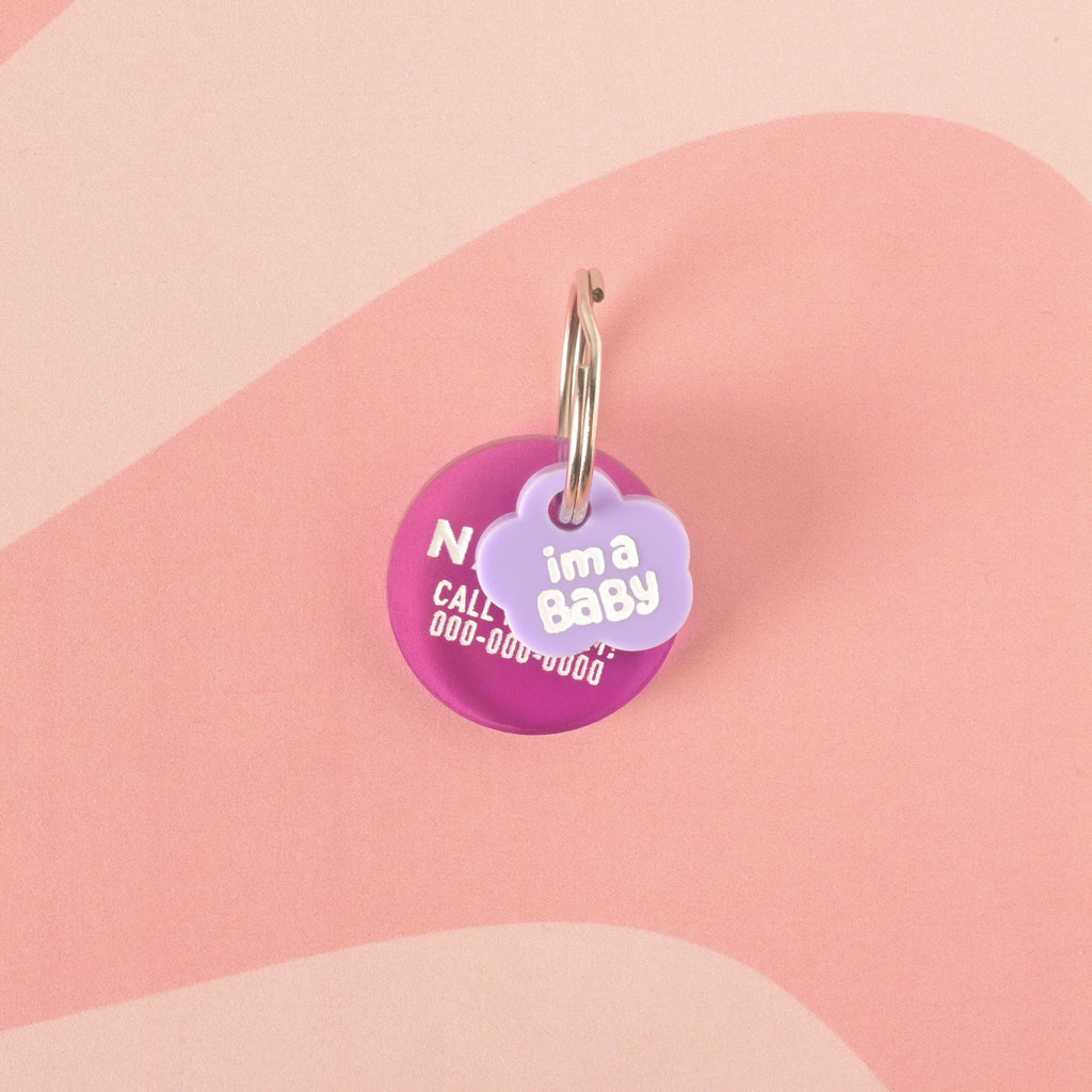 A purple cloud-shaped pet charm that says "i'm a Baby" in white letters. The charm is attached to a larger round, dark pink pet tag that has a pet's name and placeholder phone number. Both are connected to a silver ring, suitable for a collar, and are displayed on a solid pink background with a wavy design.