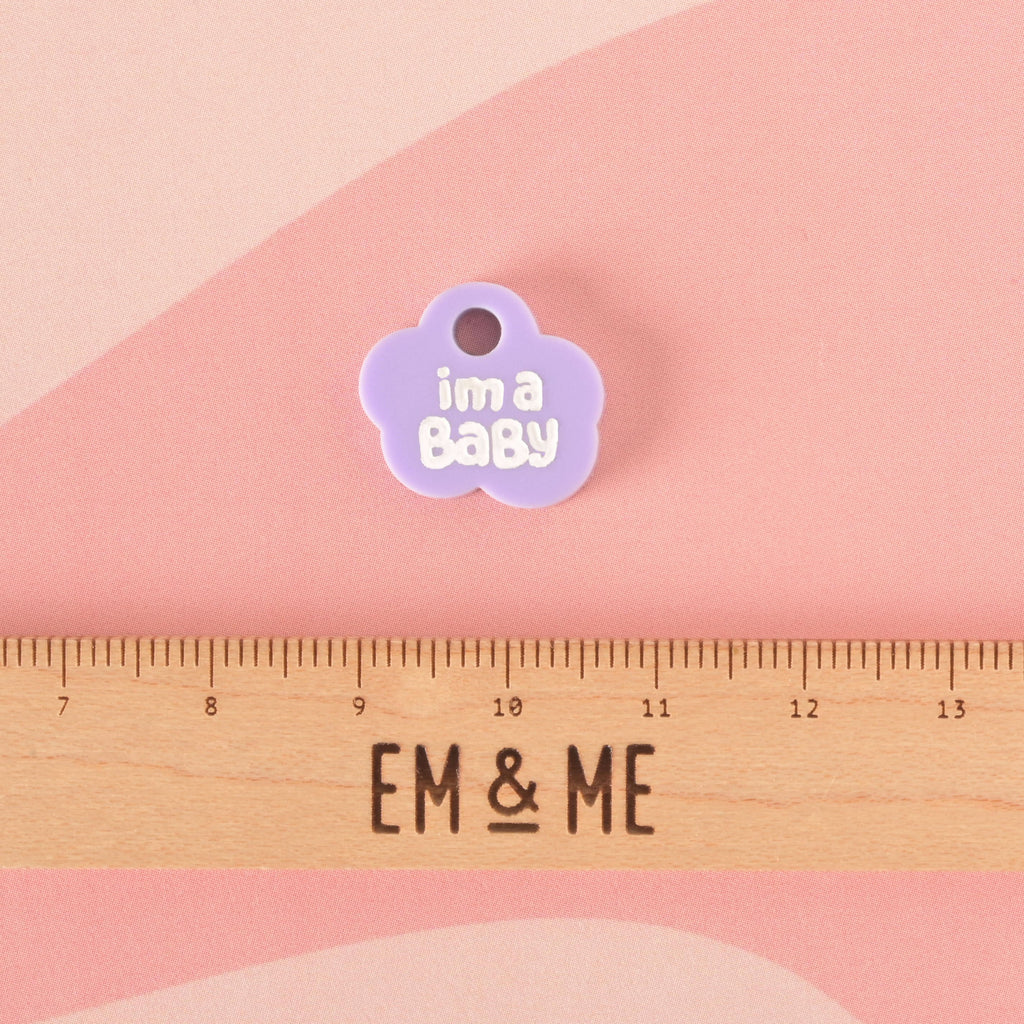 A close-up of a small, light purple cloud-shaped charm that says "i'm a Baby" in white letters. A wooden ruler with the "EM & ME" logo is placed below the charm to show its size. The charm is displayed on a light pink and light purple background.