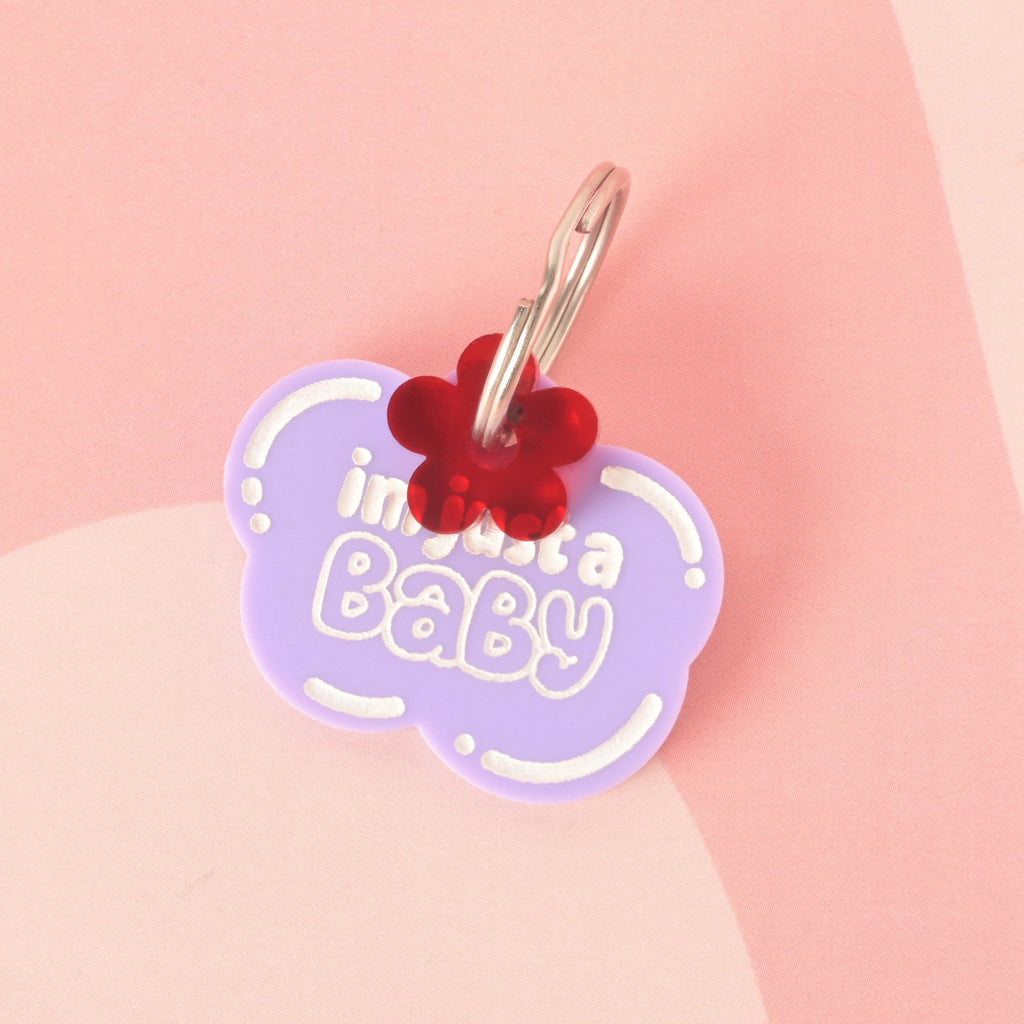A purple, cloud-shaped pet tag that says "im just a Baby" in white letters. A small red poppy flower charm is attached to the tag. Both are connected to a silver ring, perfect for a collar. The items are shown on a light pink and white checkered background.