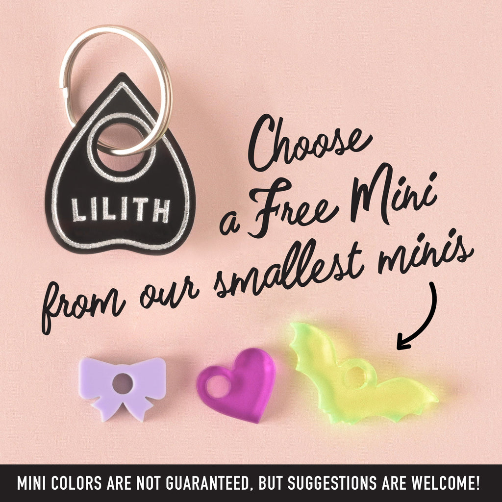 A black Ouija board planchette-shaped pet tag that says "LILITH," attached to a silver ring. The image also shows three small charms—a purple bow, a pink heart, and a translucent yellow bat. The text "Choose a Free Mini from our smallest minis" and "MINI COLORS ARE NOT GUARANTEED, BUT SUGGESTIONS ARE WELCOME!" is visible. The items are displayed on a solid pink background.