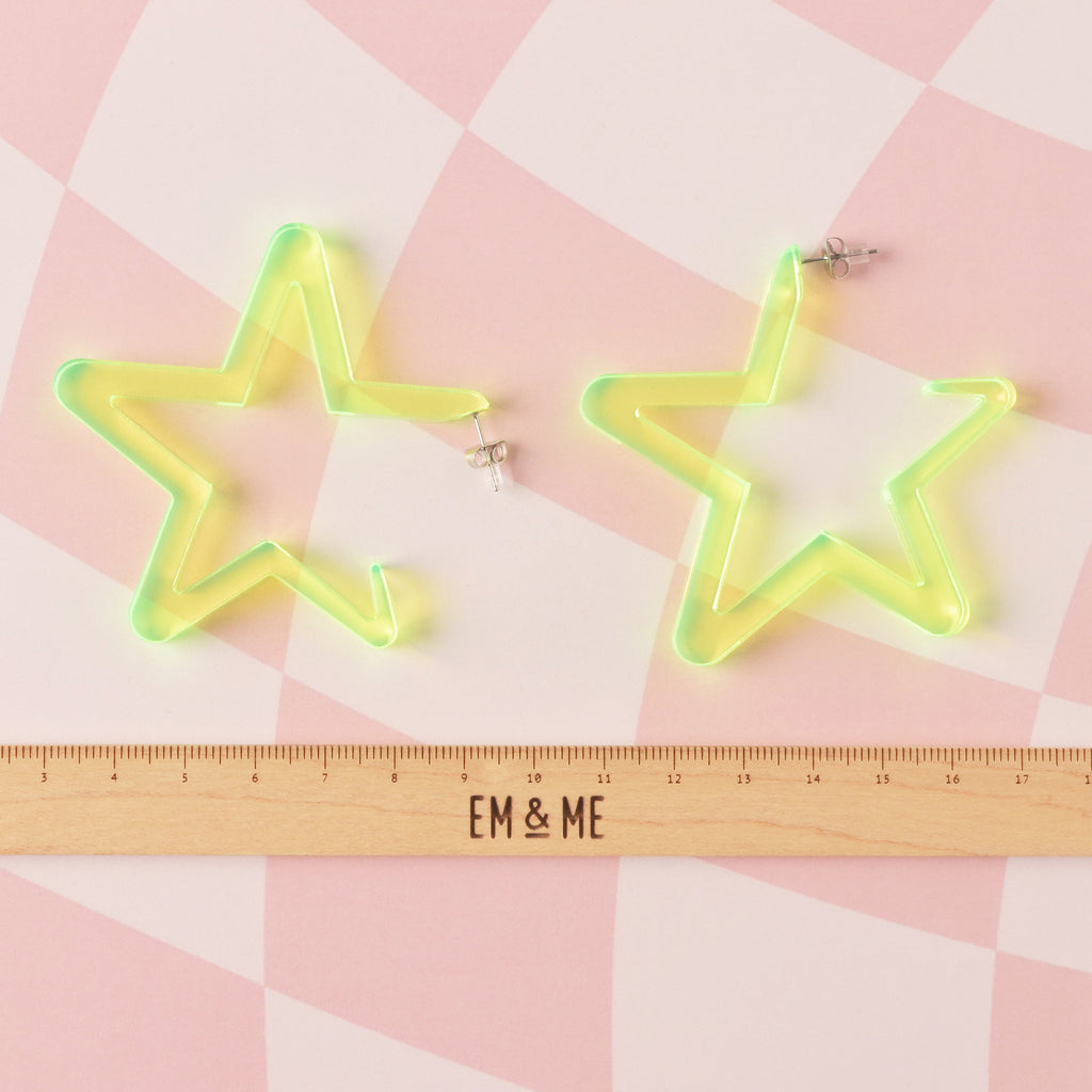 pair of oversized fluorescent neon yellow-green acrylic star hoop earrings shown above a wooden ruler with the 'EM & ME' logo, illustrating the large size of the statement jewelry.