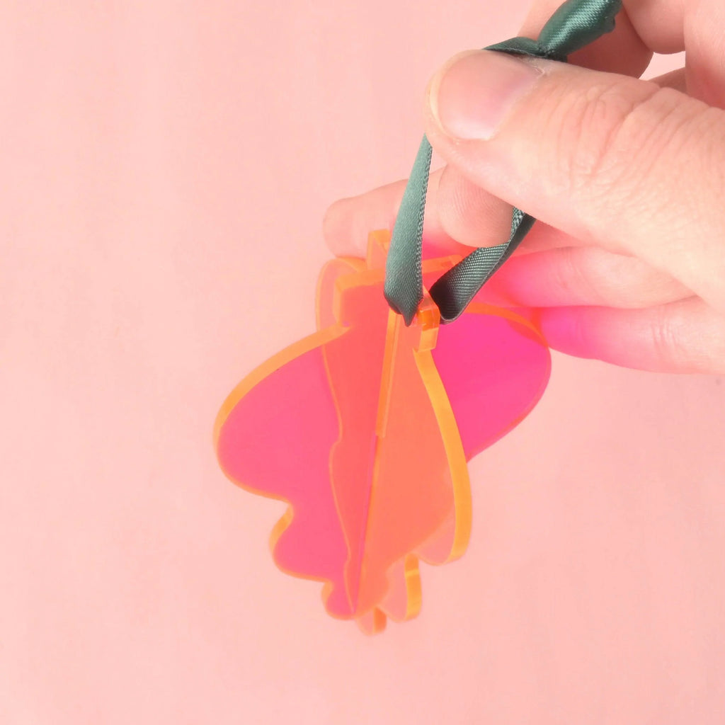 A hand holds a 3D interlocking acrylic ornament in a butterfly or abstract flourish shape. The ornament is made of layered bright pink pieces, hanging from a dark green ribbon, against a light pink background.Acrylic Icicle Christmas ornaments, bright colors and non-breakable, contemporary 70s inspired Holiday