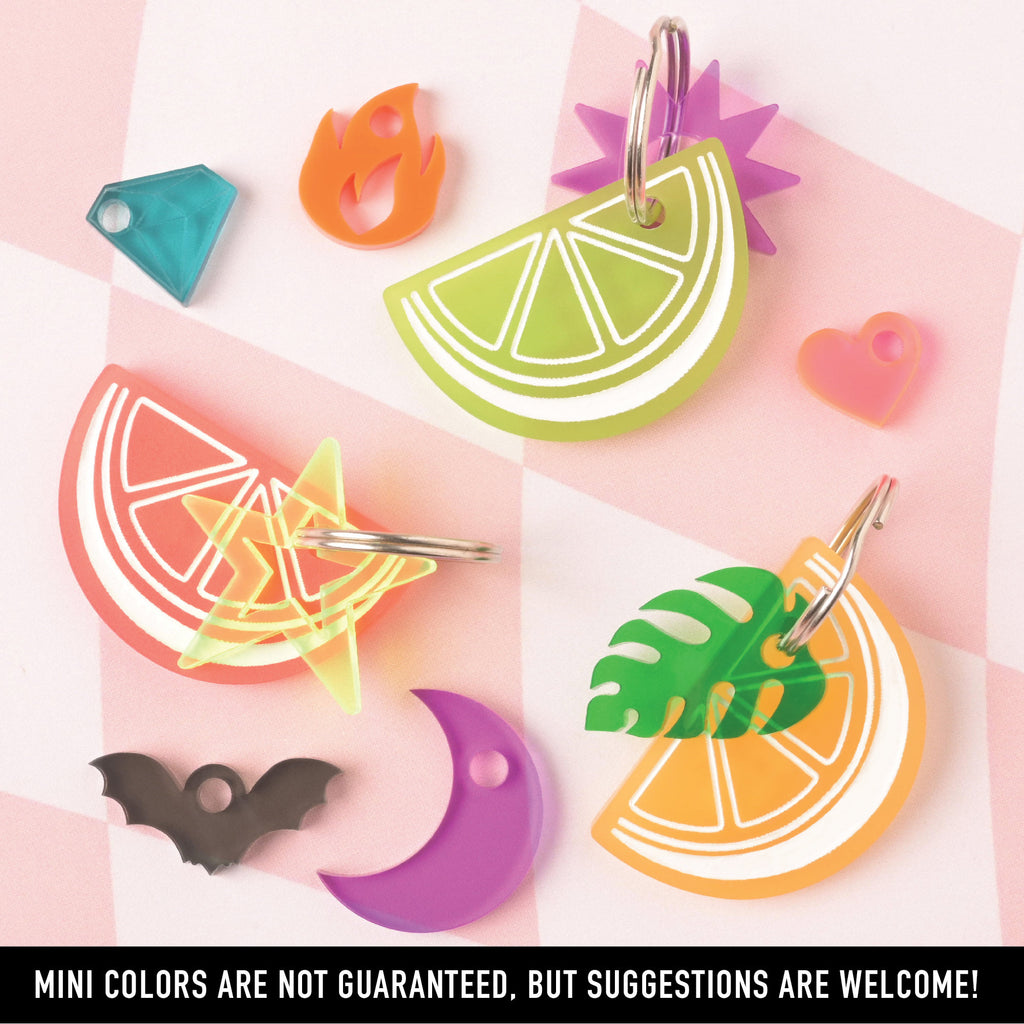 A collection of colorful acrylic mini charms: a neon lime slice, an orange slice with a tropical leaf, a grapefruit slice with a neon star, a black bat, a purple crescent moon, a turquoise gem, an orange flame, and a pink heart.