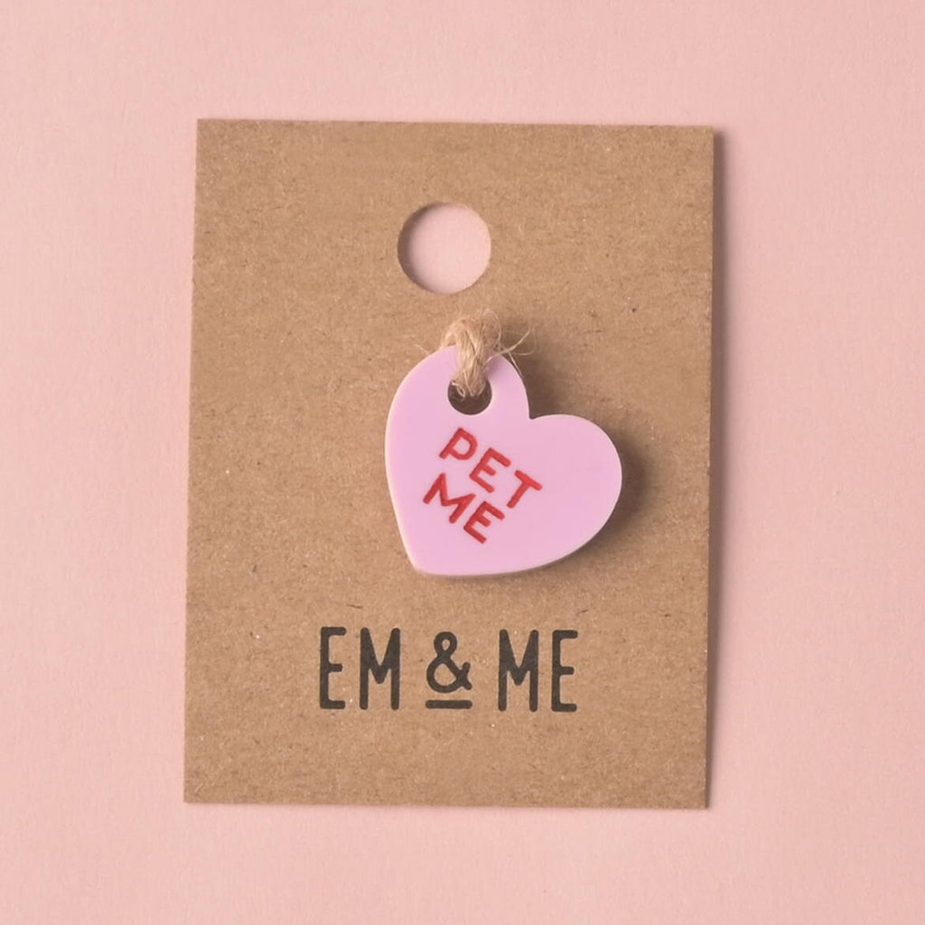 A single light purple (lilac) heart-shaped acrylic charm, engraved with the red slogan 'PET ME.' The charm is displayed on a small square kraft paper backing card with the EM & ME logo printed below it, set against a solid pink background. Small Pet Me Heart, Candy Inspired, Valentine's Day, Seasonal Mini, Tiny Accessory for Pet ID Tag, Cats and Dogs Add-on Charm