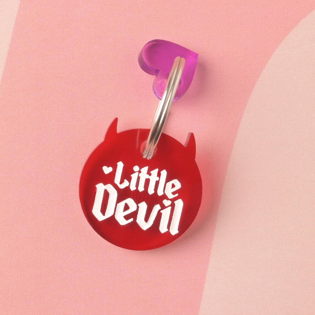Close up of red devil-shaped "little devil" pet tag with purple tiny heart mini charm on wavy pink background