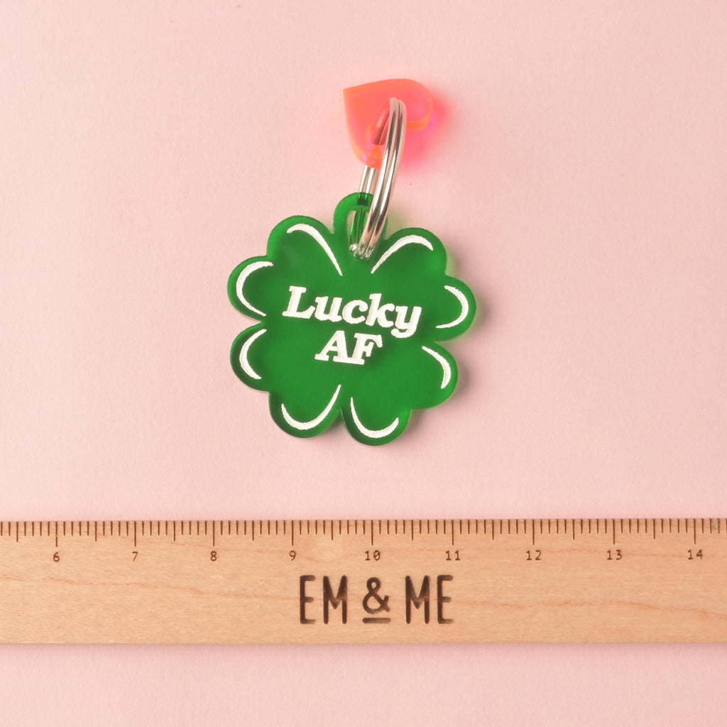 Close-up of a dark green, clover-shaped acrylic pet ID tag featuring the white, humorous slogan 'Lucky AF.' A small pink/red heart charm is attached to the silver split ring. The tag is displayed above a wooden ruler marked with the brand EM & ME, on a pink background. Lucky AF, 4 leaf Clover Personalized Pet Tag, St Patrick Cat and Dog ID Tag