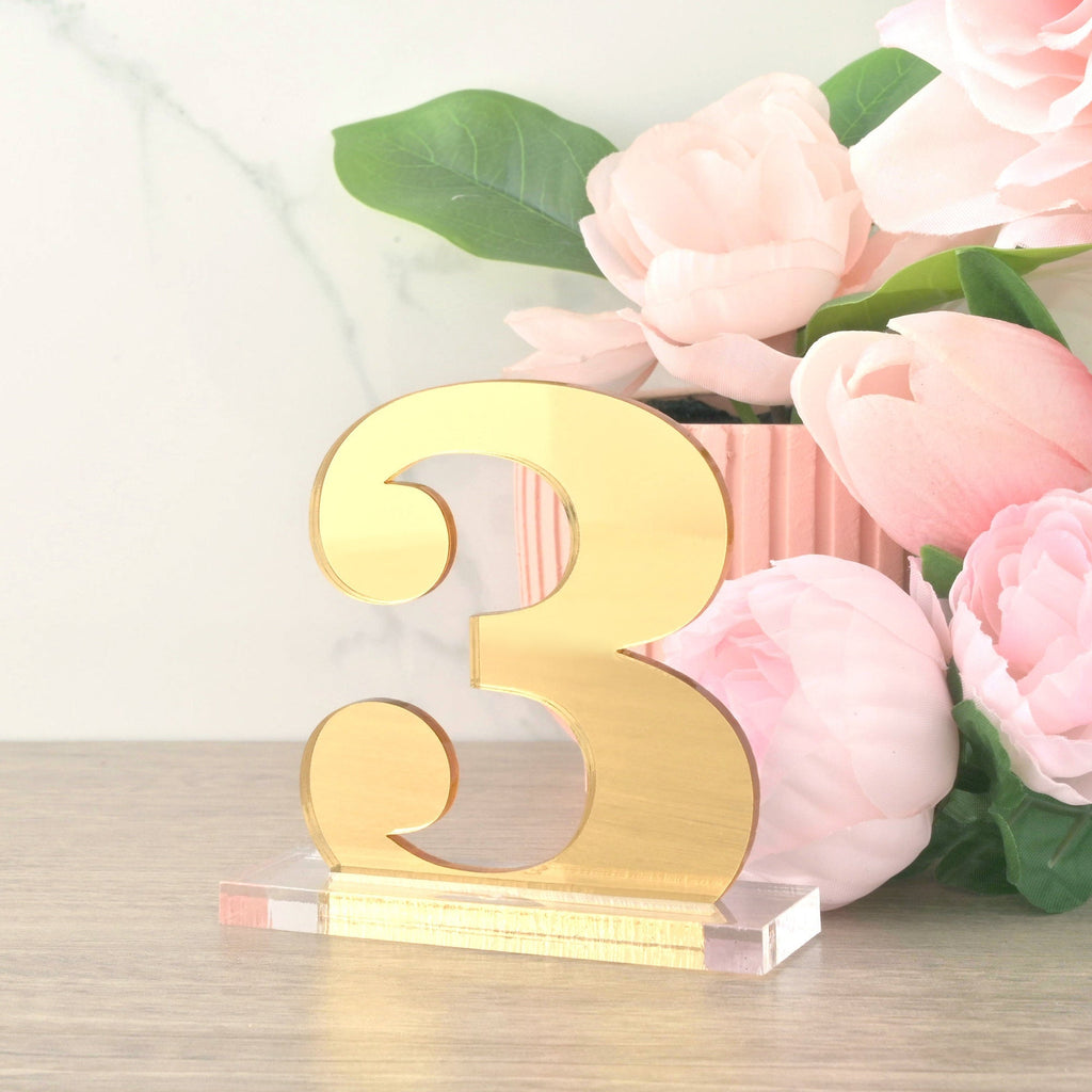 Freestanding table number '3' crafted from reflective gold mirrored acrylic in a script font, mounted on a clear acrylic base. The elegant sign is set next to soft pink flowers and a white marble background.