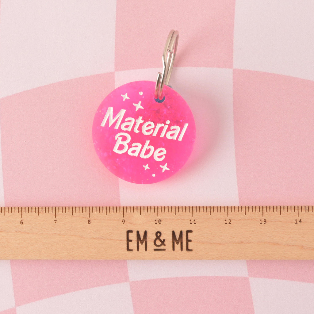  A round acrylic tag made from vibrant hot pink glitter, engraved with the white slogan "Material Babe" and small star graphics. The tag is shown above a wooden ruler with the 'EM & ME' logo for size reference.Material Babe, Barbie Inspired Personalized Pet Tag, ID Tag for Cats and Dogs, Limited Edition, Custom Name and Contact Info Engraved