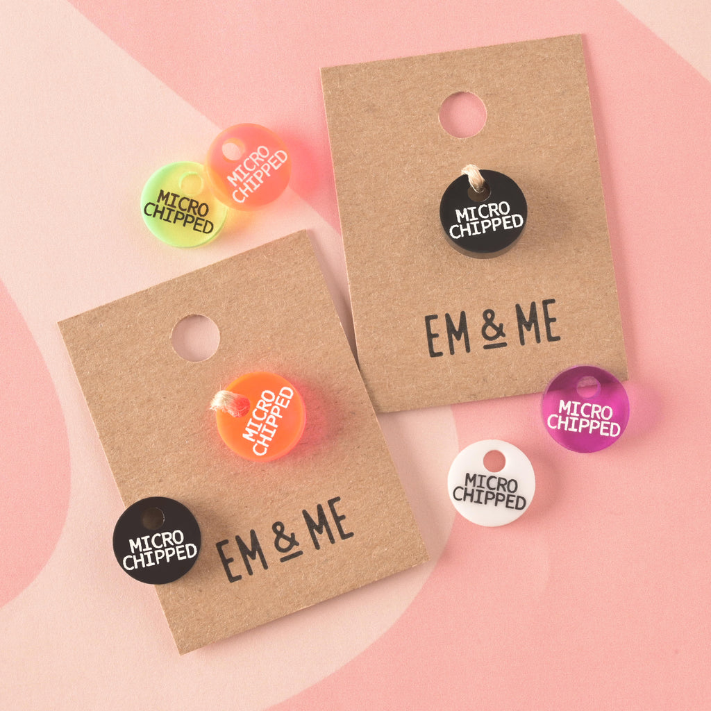 A collection of small, circular acrylic tags in various colors (black, white, neon orange, pink, yellow/green) reading "MICRO CHIPPED" on the front. Some tags are displayed on small cardboard EM & ME backing cards.
