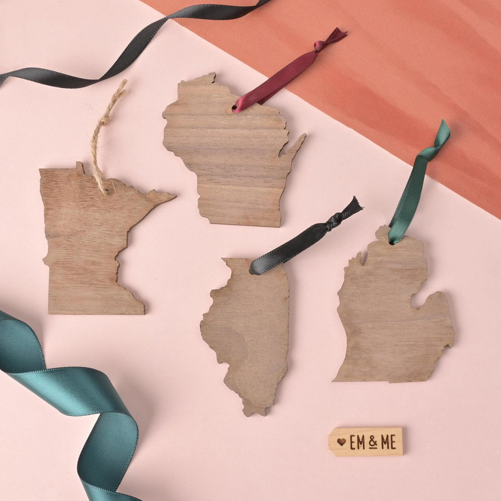 Flat lay of four wooden state-shaped ornaments representing Minnesota, Wisconsin, Illinois, and Michigan with ribbon hangers, arranged on a peach and pink background with an Em & Me wooden tag.