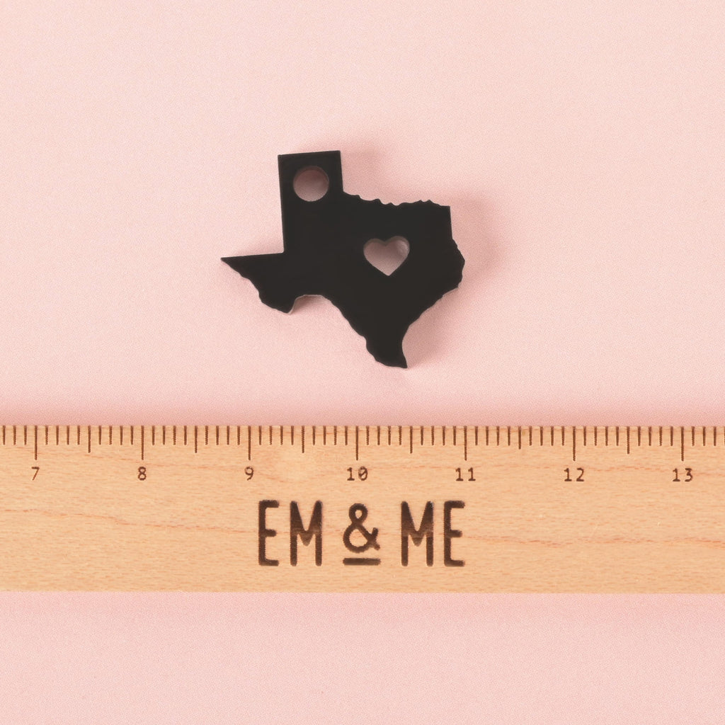 Small black acrylic Texas state-shaped charm with heart cutout, placed above a wooden EM & ME branded ruler showing size for scale, on a pink background.