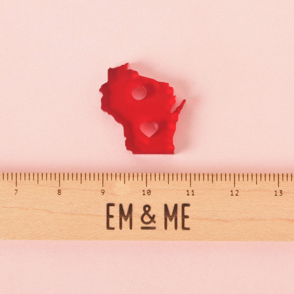 Small red acrylic Wisconsin state charm with heart cutout and hanging hole, placed above a wooden EM & ME branded ruler for size reference on a pink background.