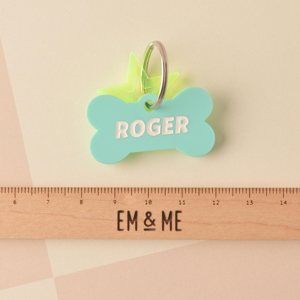 Close-up of a mint green bone-shaped acrylic pet ID tag personalized with the name 'ROGER' in white text. A translucent neon yellow-green star charm is attached to the silver split ring. The tag is displayed above a wooden ruler marked with the brand EM & ME. Bones Day, Bone shaped Dog Tag, Personalized Pet Tag, Cat and Dog ID Tag