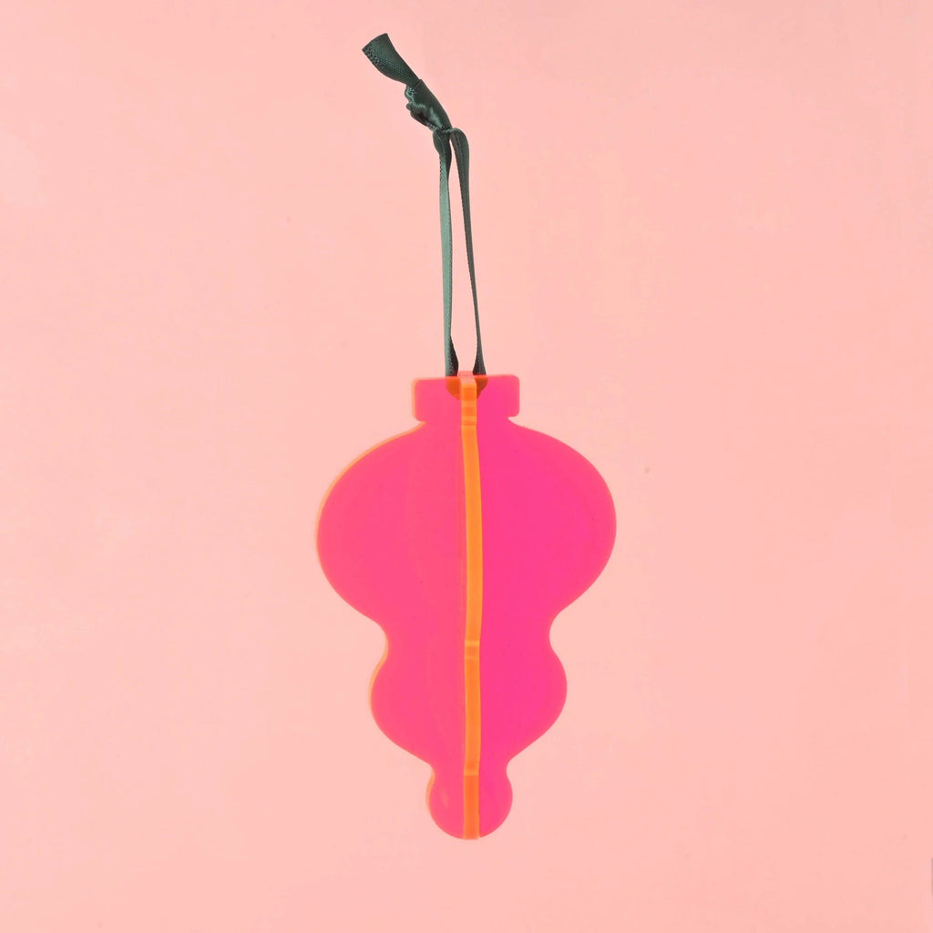 A single 3D interlocking acrylic Christmas ornament in a stylized, contoured bulb shape. It is constructed from layered bright fluorescent  pink acrylic. The ornament hangs from a dark green ribbon against a solid pink background. Acrylic Icicle Christmas ornaments, bright colors and non-breakable, contemporary 70s inspired Holiday