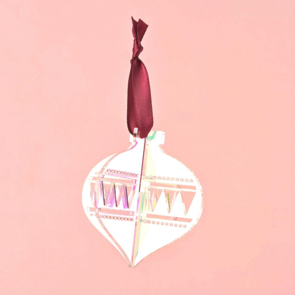 A single 3D interlocking acrylic Christmas ornament in a clear or iridescent finish, featuring engraved geometric patterns. The ornament is hanging from a dark red/burgundy satin ribbon against a light pink background. Acrylic Christmas ornaments, bright colors and non-breakable