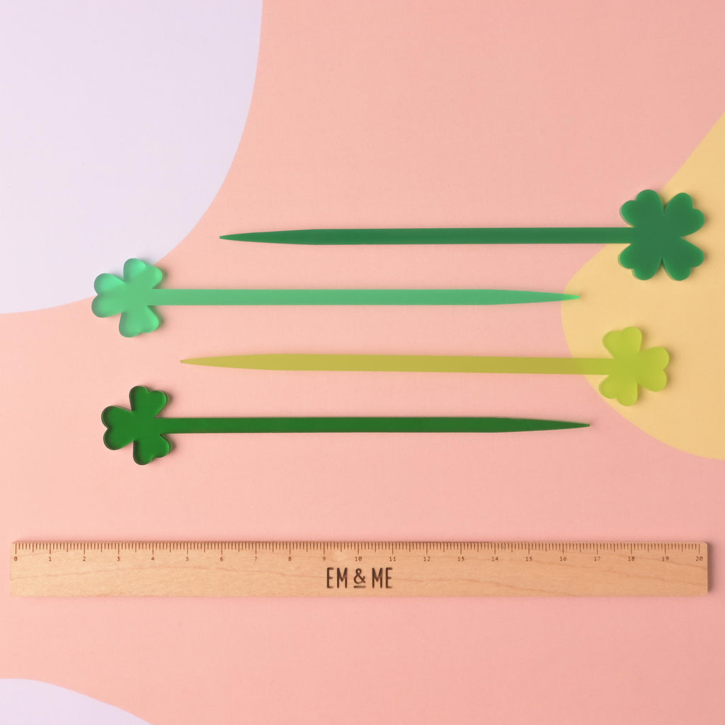 Four acrylic stir sticks with clover-shaped tips, laid out horizontally in various shades of green, including dark, medium, and lime green. They are displayed above a wooden ruler marked with the brand EM & ME, on a color-blocked background.