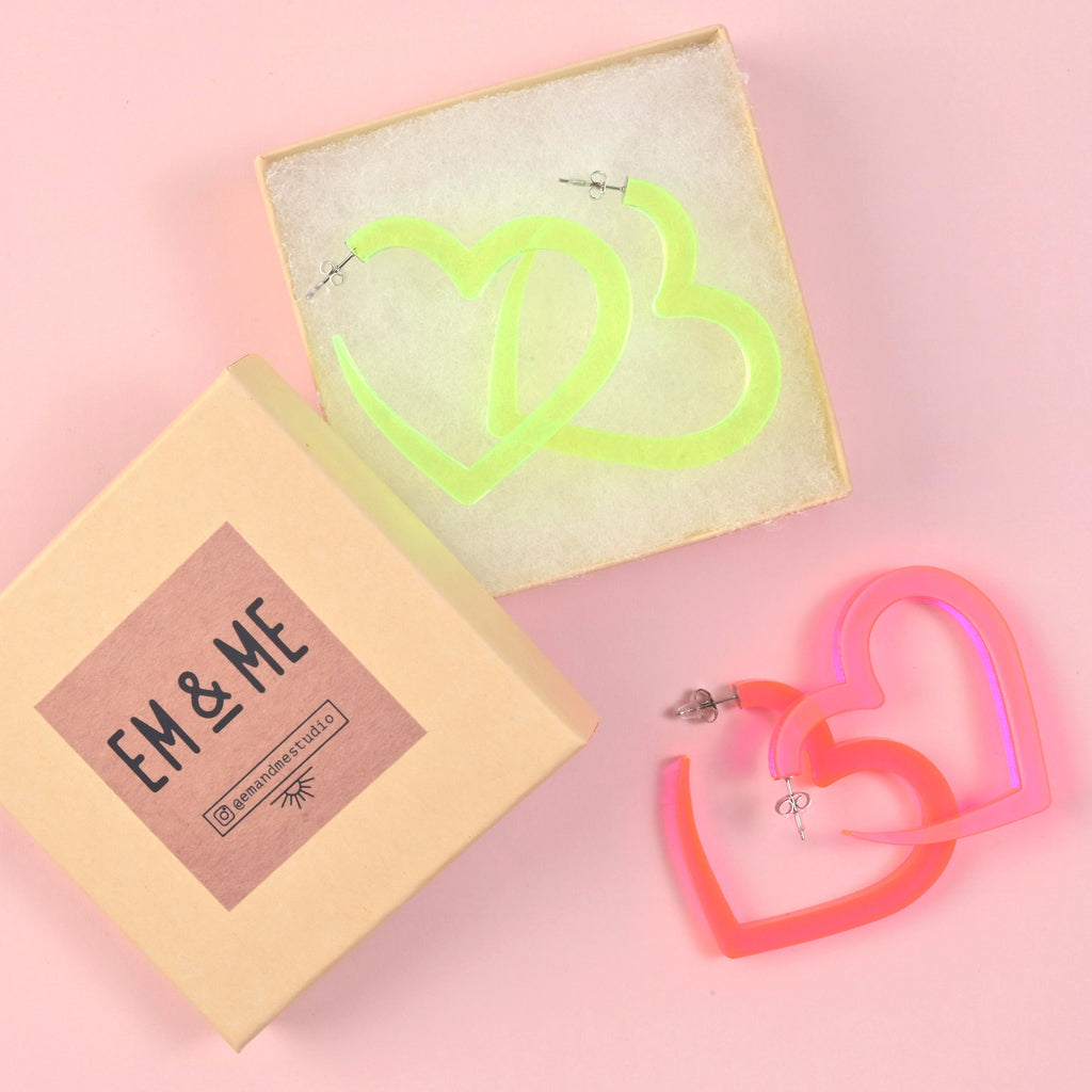 A pair of oversized fluorescent neon green acrylic heart hoop earrings sits in an open gift box. A second pair of pink/orange iridescent heart hoops is shown outside the box. The lid is branded with the 'EM & ME' logo.