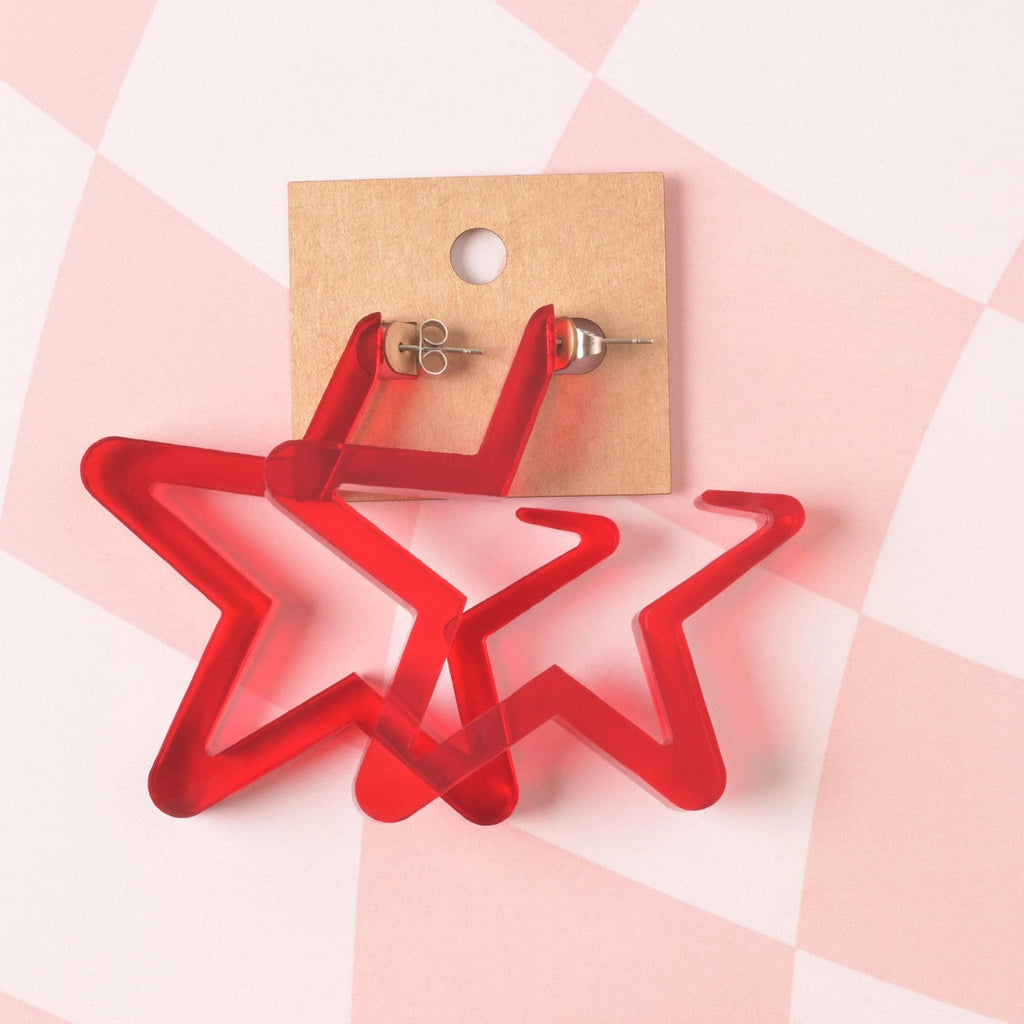 A pair of oversized, bright red translucent acrylic star hoop earrings displayed on a brown kraft jewelry card, set against a pink and white checkered background.