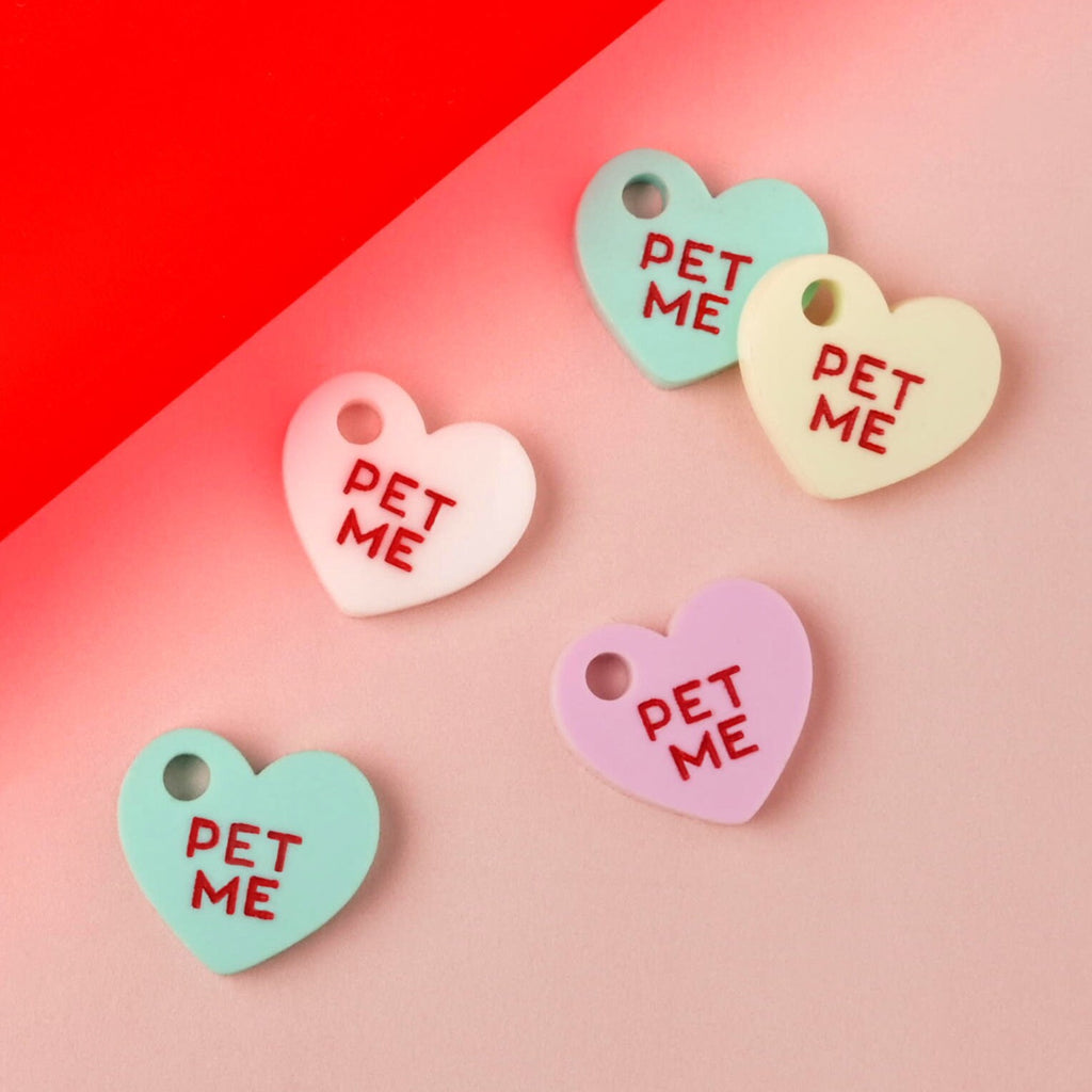 Five small, heart-shaped acrylic mini-charms, each engraved with the red slogan 'PET ME.' The charms are in assorted pastel colors: white, mint green, light yellow, and lilac purple. They are displayed on a pink and red color-blocked background. Small Pet Me Heart, Candy Inspired, Valentine's Day, Seasonal Mini, Tiny Accessory for Pet ID Tag, Cats and Dogs Add-on Charm